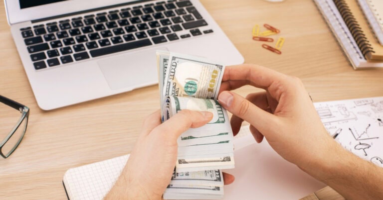 male hands holding a wad of cash with a laptop and stationary - how to make money with a laptop