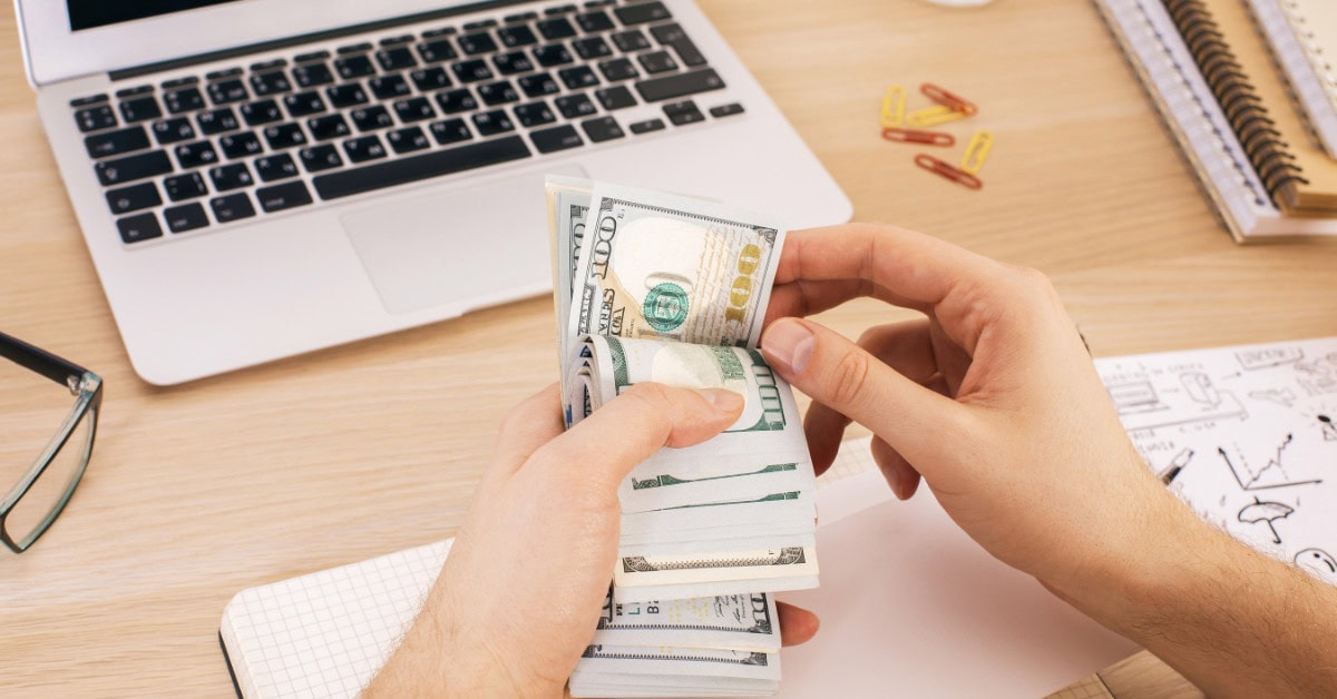 male hands holding a wad of cash with a laptop and stationary - how to make money with a laptop